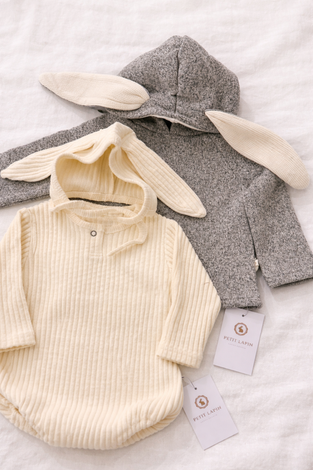 Two children's set hoodie and bodysuit with bunny ears, hoodie in gray and bodysuit in cream, on a light background.