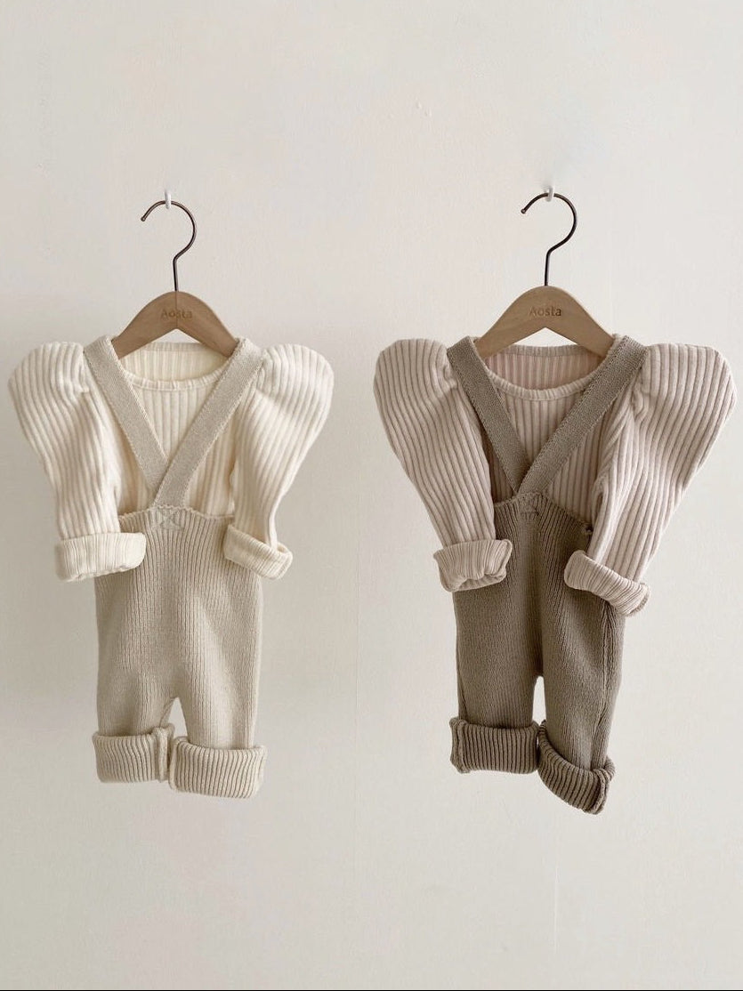 Two sets of baby rompers with suspenders on wooden hangers against a plain background.